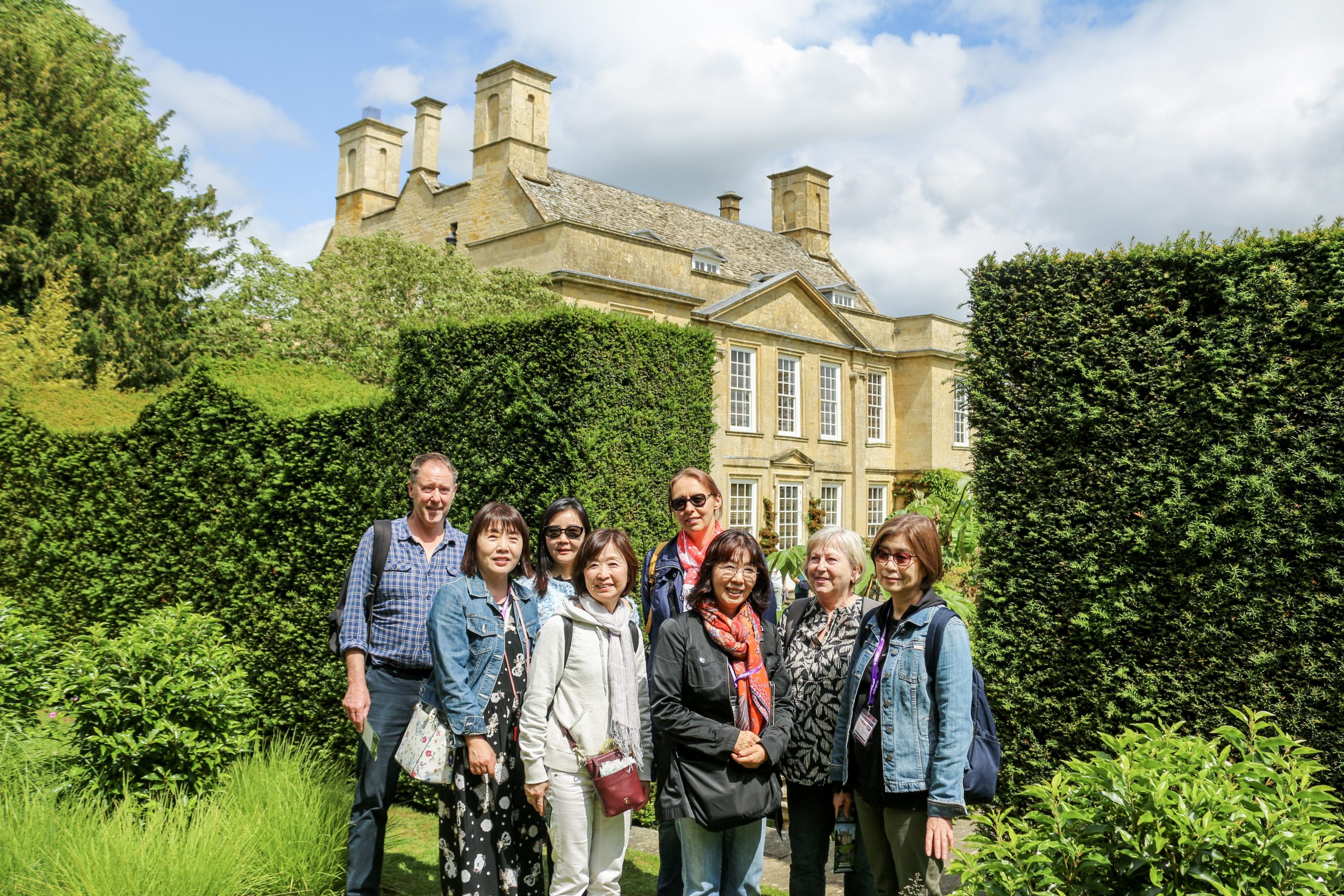 A Gardens and Culture class of adult students aged 50 and over standing with their teacher in front of a Cotswold stone manor house, surrounded by clipped hedges and summer planting